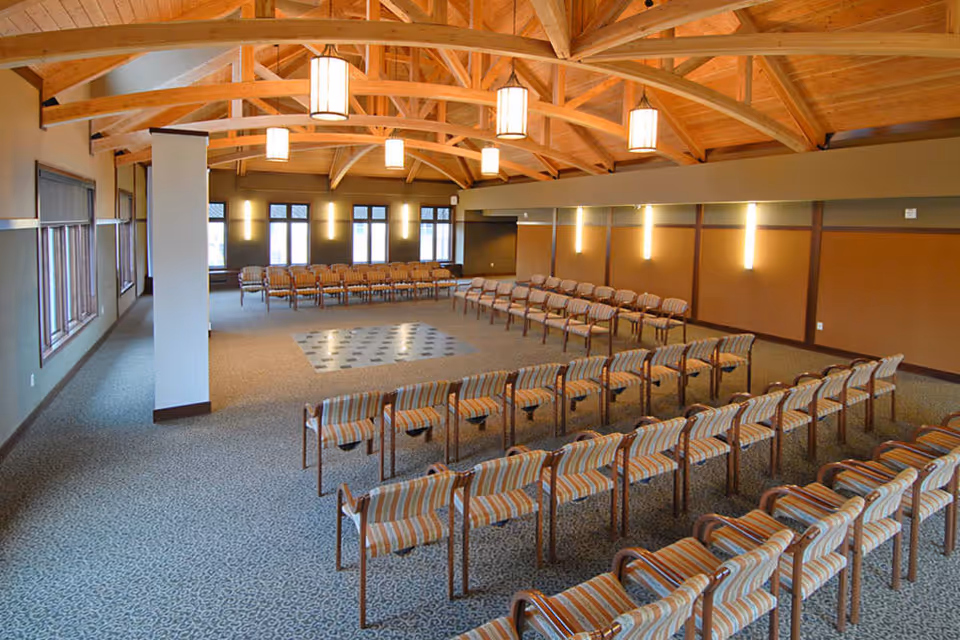 Large meeting or event room with rows of striped chairs arranged in a U-shape around a central open space. The room features a high wooden ceiling with exposed beams and hanging pendant lights. Windows line one side of the room, and the walls are decorated with vertical light fixtures.