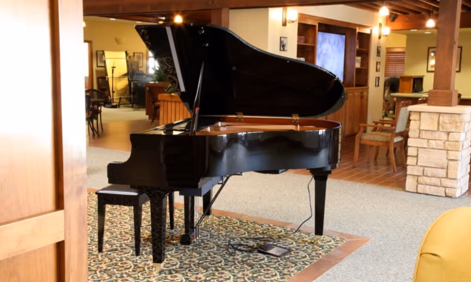 A black grand piano with an open lid sits on a patterned rug in a warmly lit room with wooden beams and furniture. The room has a cozy and inviting atmosphere with chairs, a stone pillar, and built-in shelves in the background.