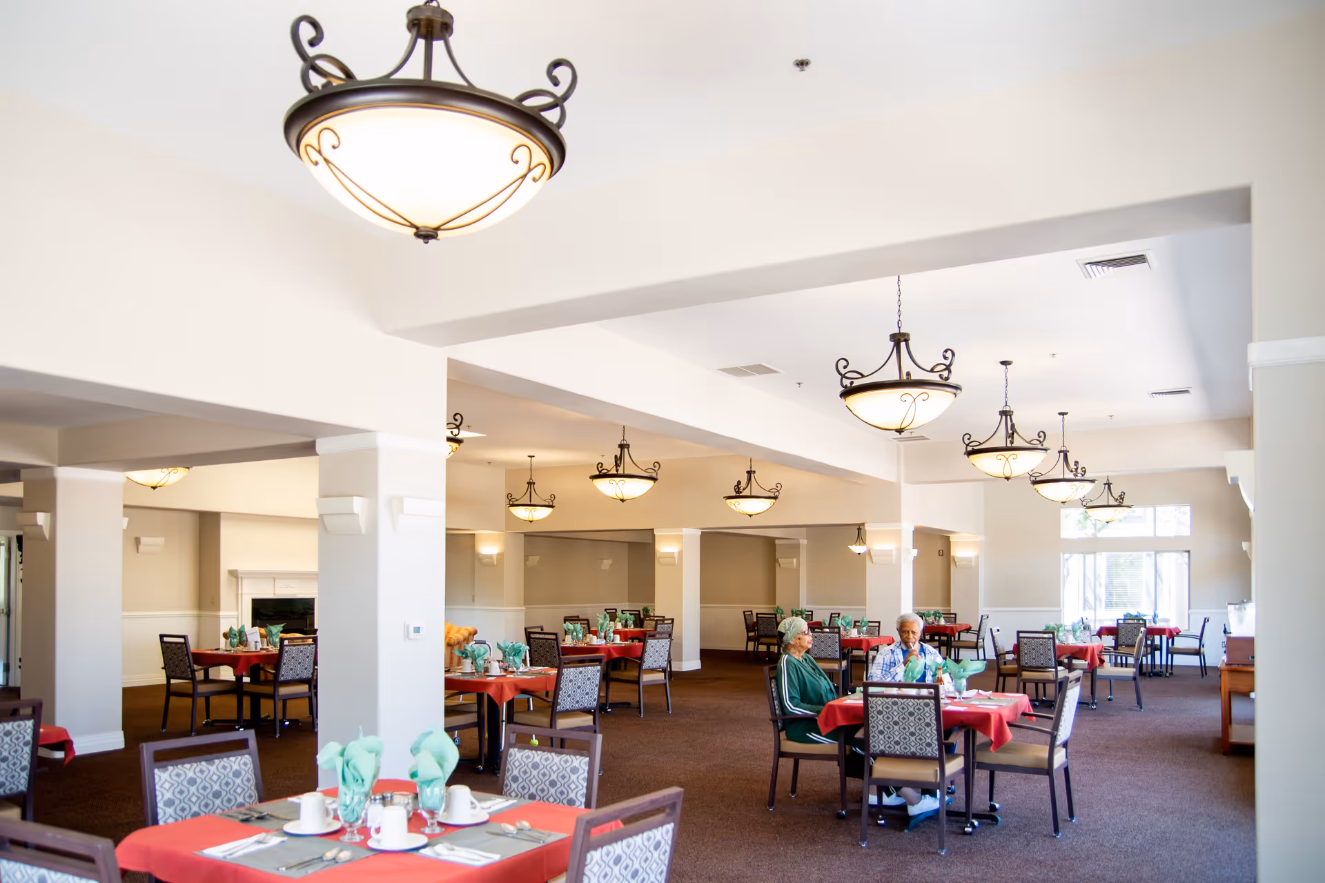 A spacious dining room in a senior living facility with multiple tables covered in red tablecloths and set with green napkins, cups, and utensils. Two elderly women are seated at one of the tables, engaged in conversation. The room features elegant hanging light fixtures and large windows allowing natural light to fill the space.