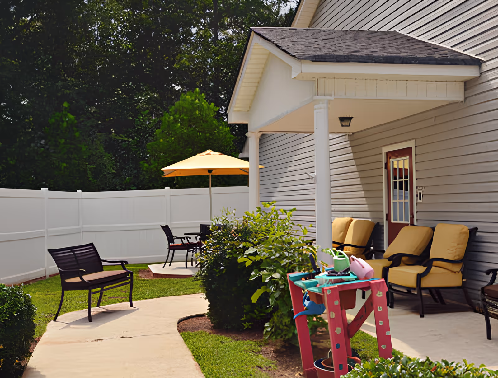Outdoor patio area at Brookside Stone Mountain with a concrete walkway, metal bench, yellow cushioned chairs under a covered porch, a table with a yellow umbrella, and green bushes along the side of the building.