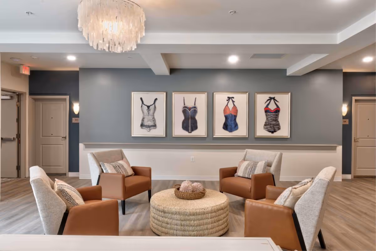 A seating area in a senior living common room with four armchairs around a woven ottoman and framed swimsuit artwork on a blue-gray wall.