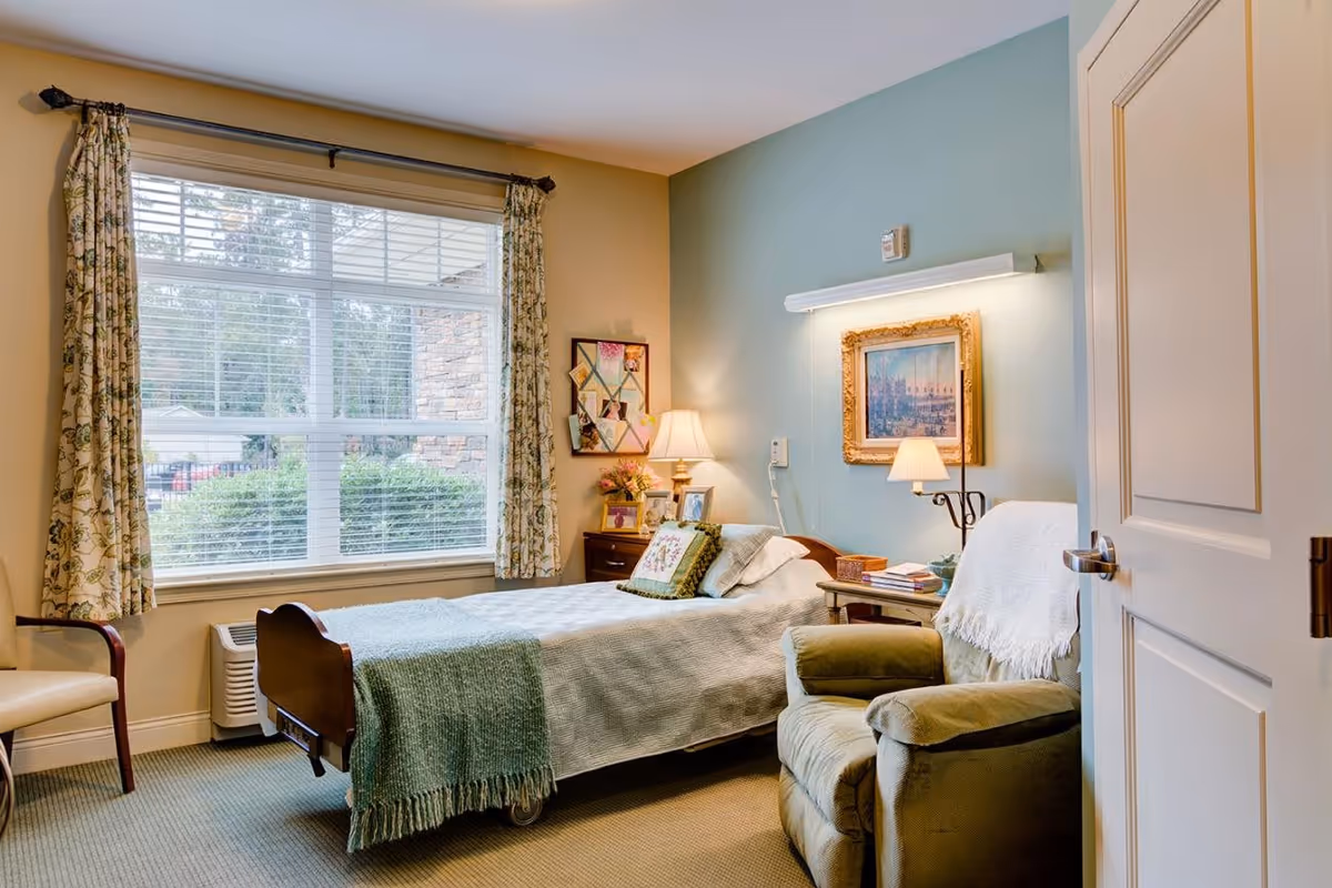 A cozy bedroom in a senior living facility with a single bed covered in a white quilt and a green throw blanket. Next to the bed is a wooden nightstand with a lamp, picture frames, and flowers. A comfortable green armchair with a white throw is positioned near the bed. The room has a large window with floral curtains letting in natural light, and a framed painting is hung on the light blue wall above the bed.