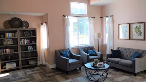 A cozy living room area with a gray sofa and two matching armchairs arranged around a round coffee table. The room has light pink walls, three windows with white curtains, a tall floor lamp, two framed botanical prints on the wall, and a bookshelf filled with books and decorative items.