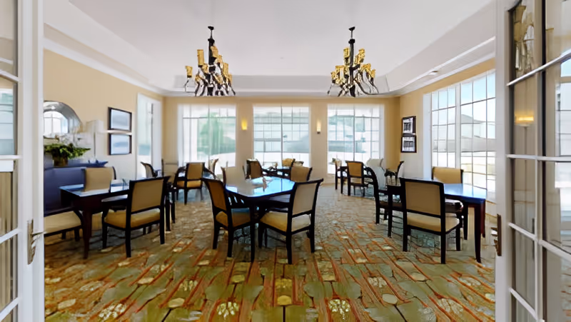 A bright and spacious dining room with multiple tables and chairs arranged neatly. The room features large windows allowing natural light to fill the space, two elegant chandeliers hanging from the ceiling, and a patterned carpet. The walls are painted in a soft yellow tone, and there are framed pictures and a bookshelf on the walls.