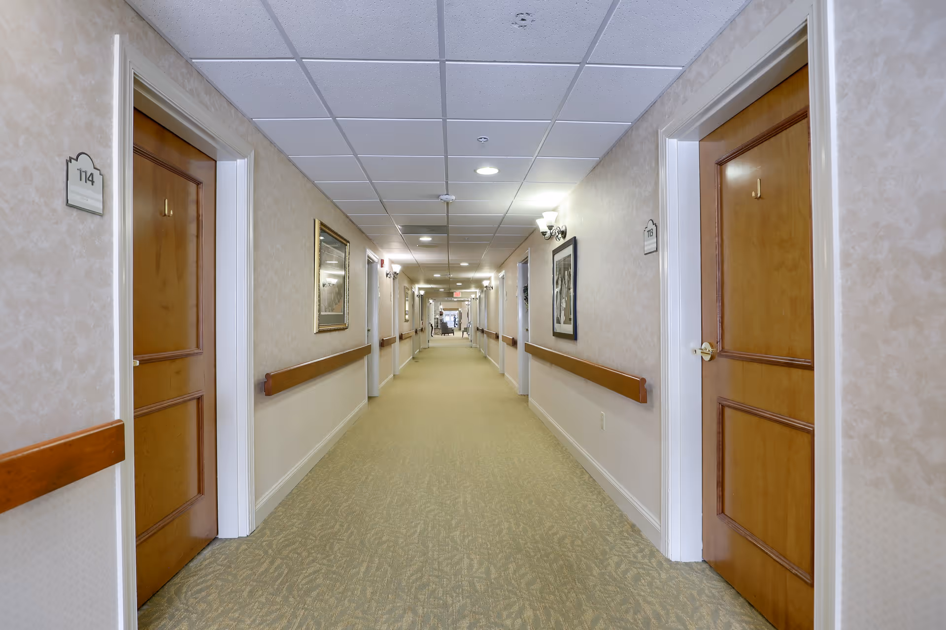 Long hallway in a senior living facility with beige walls, carpeted floor, wooden handrails on both sides, and multiple closed wooden doors with room numbers. The ceiling has recessed lighting and a drop ceiling with white tiles. There are framed pictures on the walls and wall-mounted light fixtures.