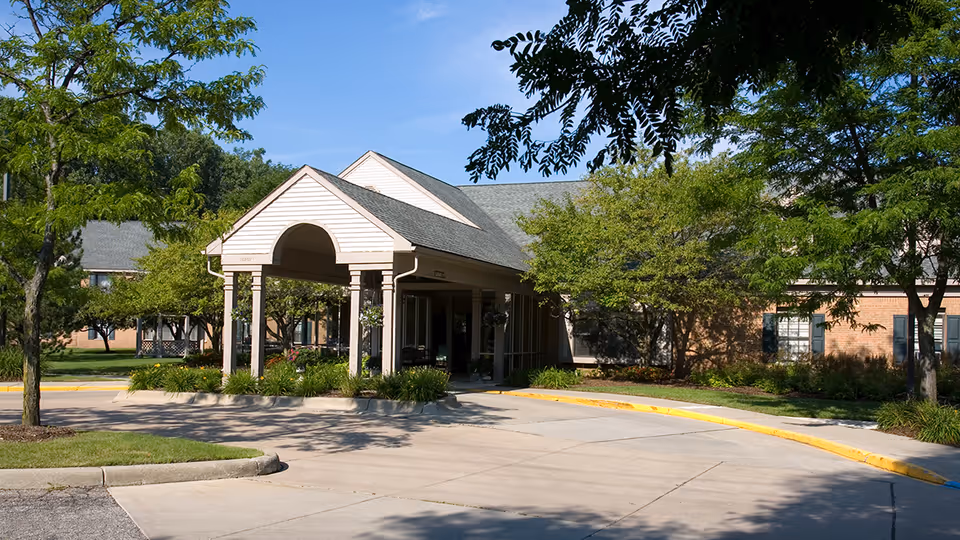 Exterior view of a senior living facility entrance with a covered drop-off area, surrounded by trees and landscaping under a clear blue sky.