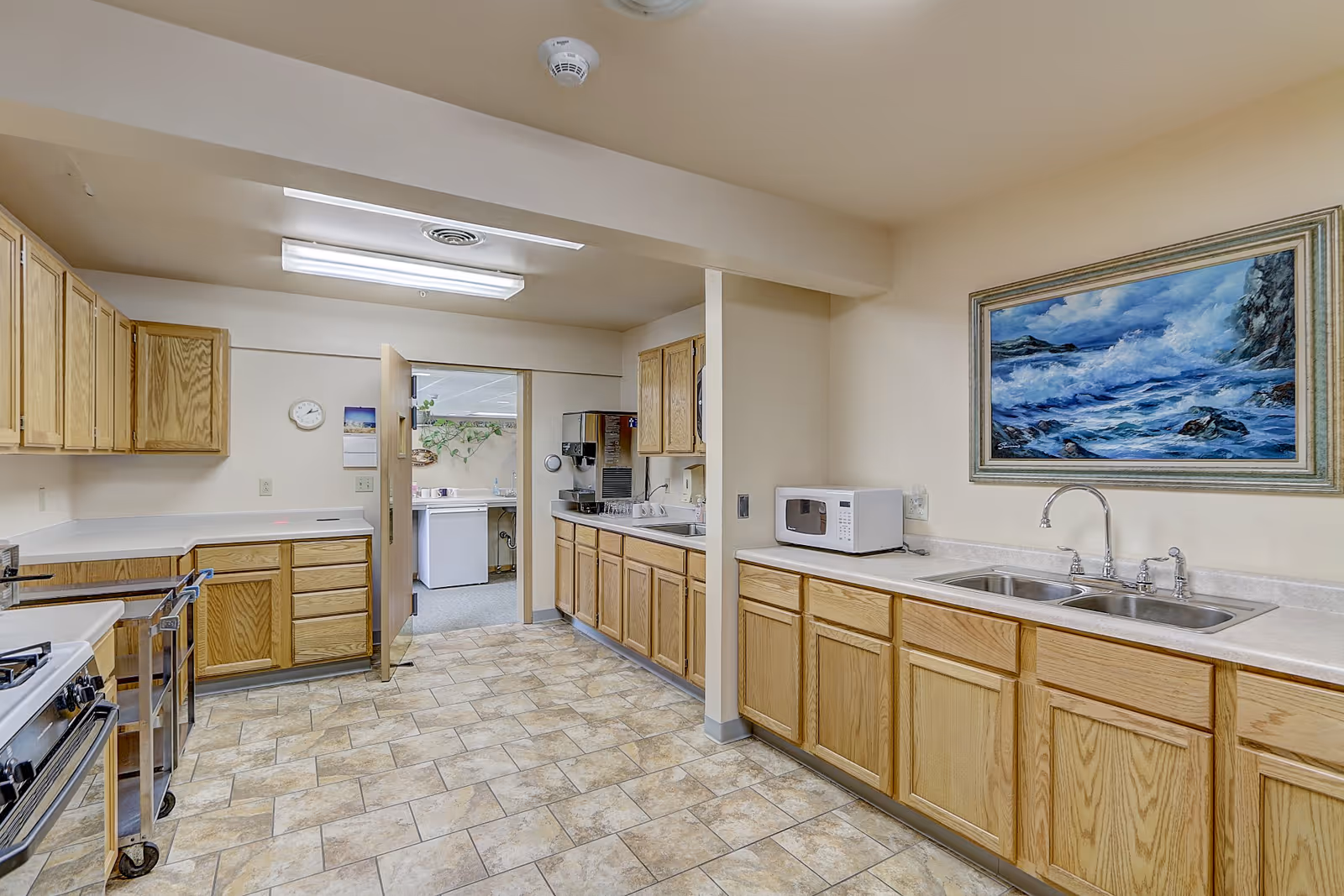 A spacious kitchen with light wood cabinets and beige countertops. The floor is tiled with a beige and brown pattern. There is a double sink with a silver faucet on the right side, a microwave on the counter, and a stove on the left. A painting of a seascape with waves crashing on rocks hangs on the wall above the sink. The kitchen opens into another room with a door that is partially open, revealing a washing machine and some plants on the far wall.