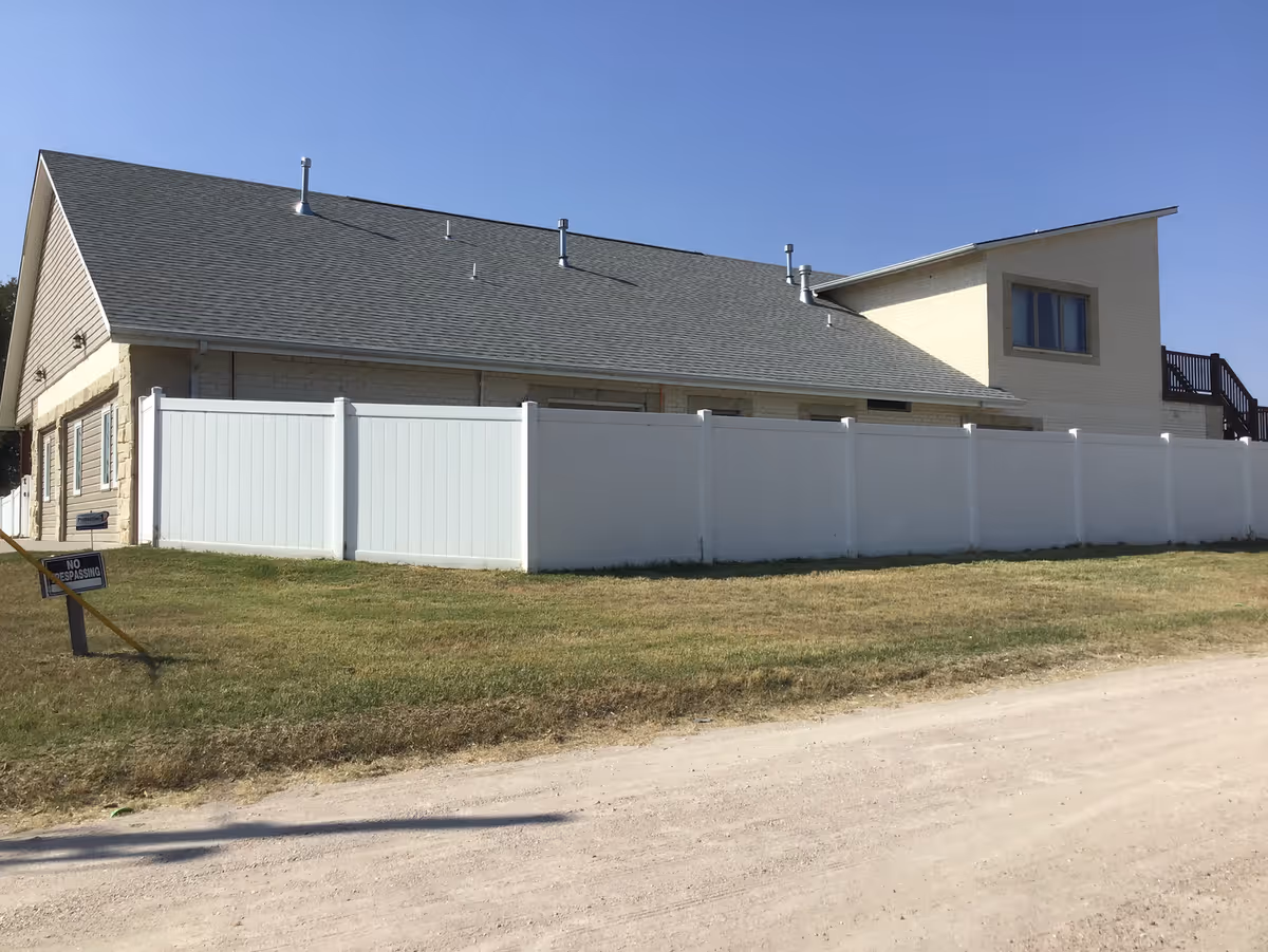 Exterior side view of a single-story building with a gray shingled roof and light-colored brick walls, surrounded by a white privacy fence. There is a grassy area and a dirt road in the foreground under a clear blue sky.