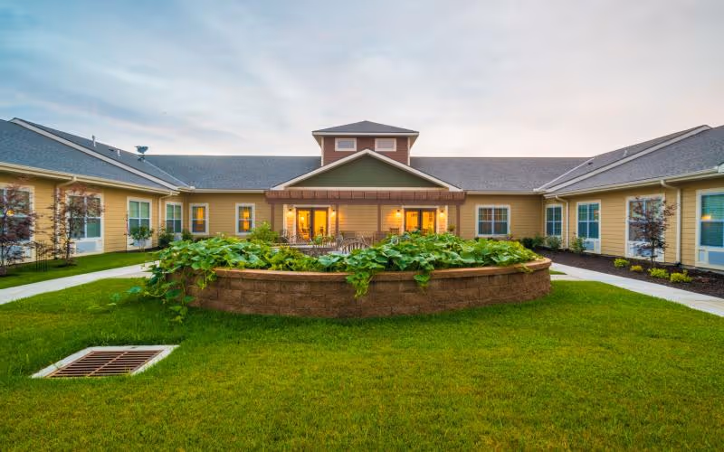 View of a courtyard garden area with green grass and a raised circular planter filled with plants, surrounded by a single-story building with beige siding and multiple windows. The building has a covered patio area with outdoor seating and warm lighting inside.
