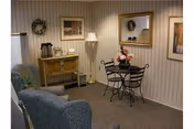 Small cozy sitting area with two upholstered armchairs, a round wrought-iron table with chairs and a floral centerpiece, a sideboard with coffee urns, a floor lamp, mirror and framed artwork on striped wallpaper.
