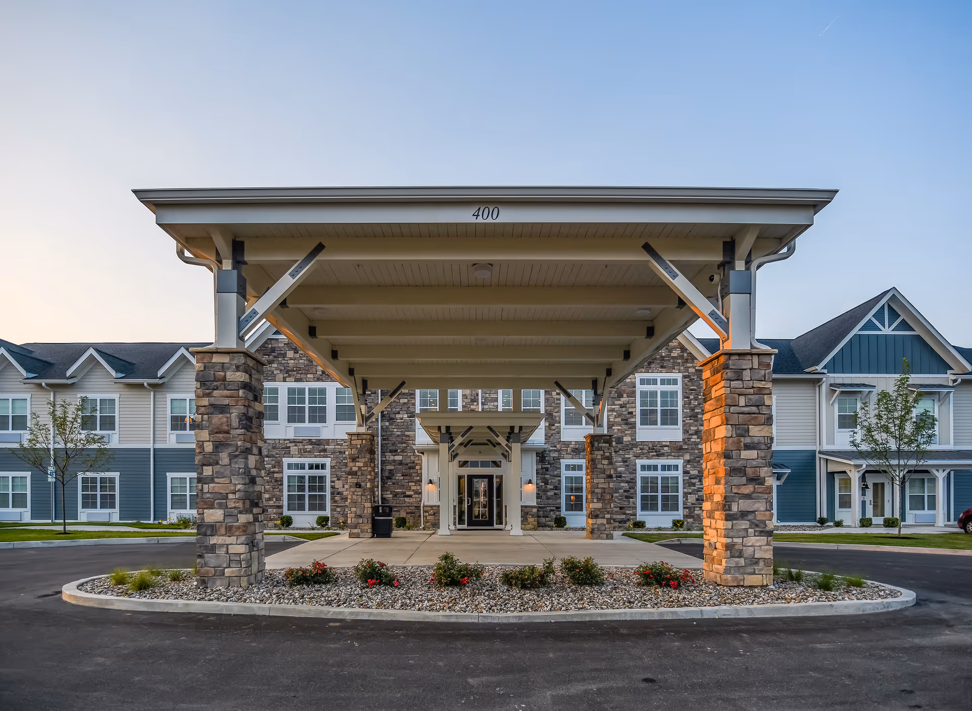 Front exterior view of Traditions at Hunter Station, showing a covered entrance supported by stone pillars, with a landscaped area featuring small bushes and flowers in front. The building has multiple windows and a combination of stone and siding on the facade.