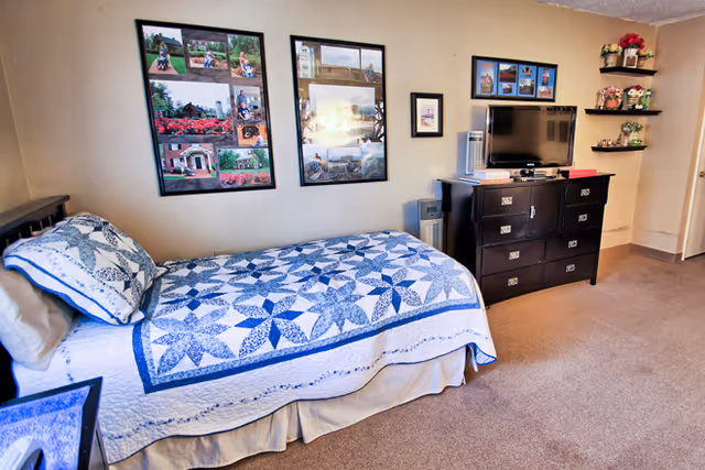 A neatly made single bed with a blue and white patterned quilt and matching pillow in a senior living facility room. Above the bed are three framed photo collages on the wall. Across from the bed is a dark wooden dresser with a flat-screen TV on top, a small fan, and some decorative items. To the right, there are two small wall shelves with flower arrangements and figurines. The room has beige walls and carpeted flooring.