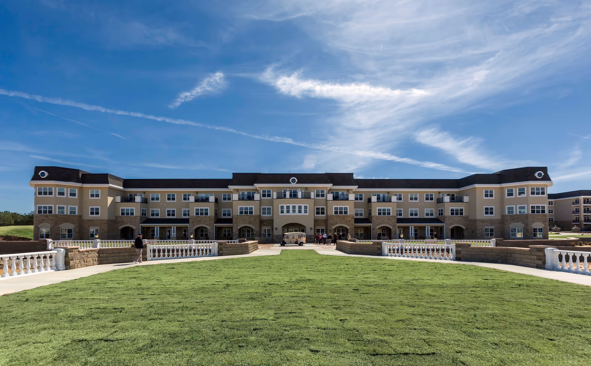 Wide exterior view of a large, multi-story senior living facility building with beige and brown tones, surrounded by a well-maintained green lawn and a clear blue sky with wispy clouds.