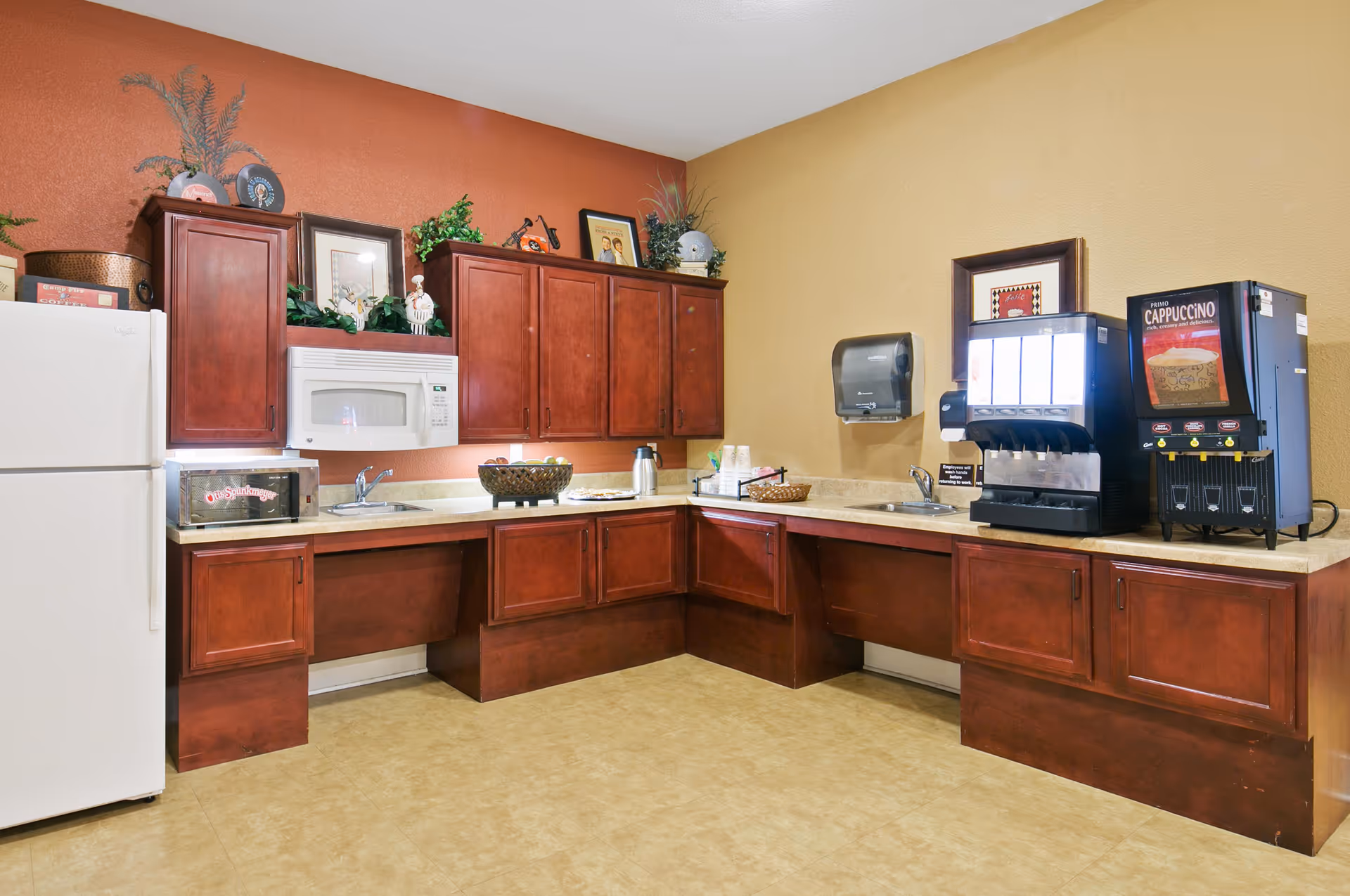 Communal kitchenette with cherry wood cabinets, countertops, sinks, a refrigerator, microwave and drink dispensers.