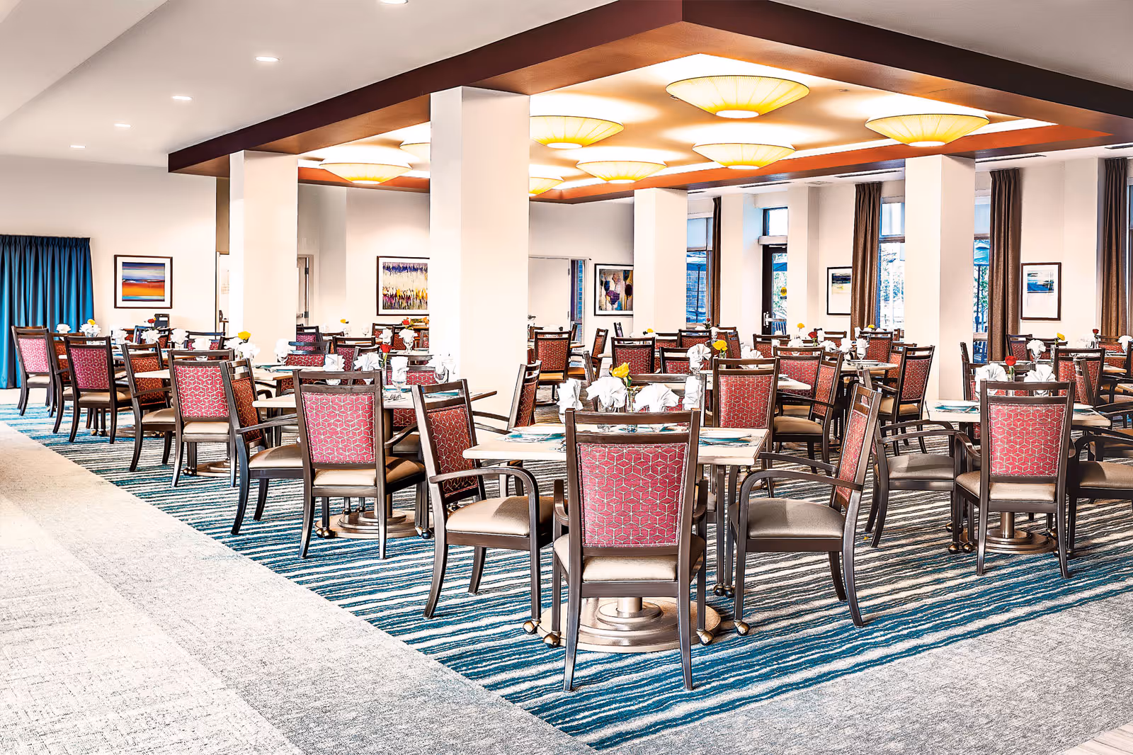 Spacious dining room with multiple tables set for service and red-upholstered chairs under warm ceiling lights.