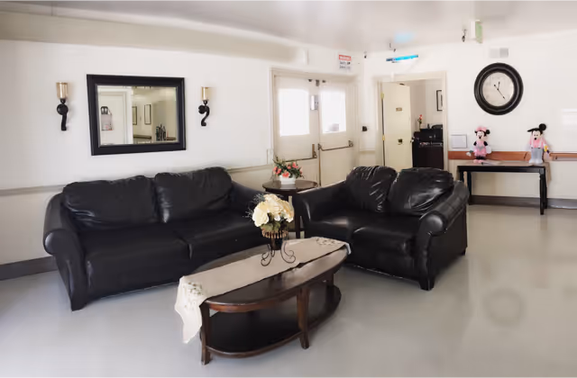 A common area with two black leather sofas arranged around a wooden coffee table with a floral centerpiece. A large mirror and two wall sconces are mounted on the wall behind one sofa. In the background, there is a door with windows, a small table with two Minnie Mouse plush toys, and a large round wall clock.