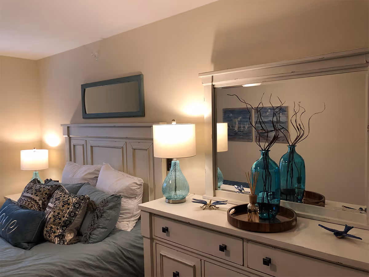 A cozy bedroom featuring a bed with multiple pillows and a headboard, two bedside lamps with blue glass bases, a dresser with a large mirror above it, and decorative blue glass vases with twigs on the dresser.