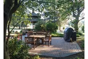 Outdoor patio area with round wooden tables and chairs on a brick-paved surface, surrounded by trees and greenery, with a covered grill and a gazebo in the background.