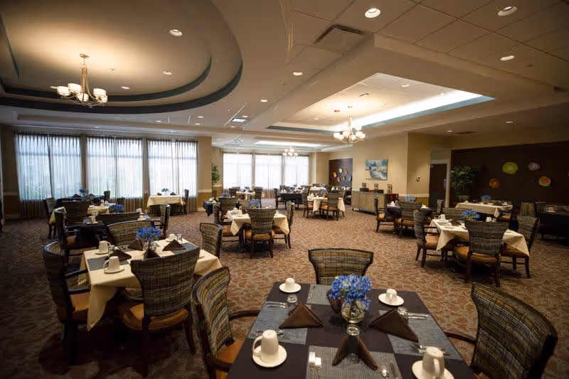 A spacious dining room with multiple tables set for meals, each table covered with tablecloths, napkins, cups, and small flower arrangements. The room has large windows with sheer curtains allowing natural light, chandeliers hanging from the ceiling, and carpeted floors.