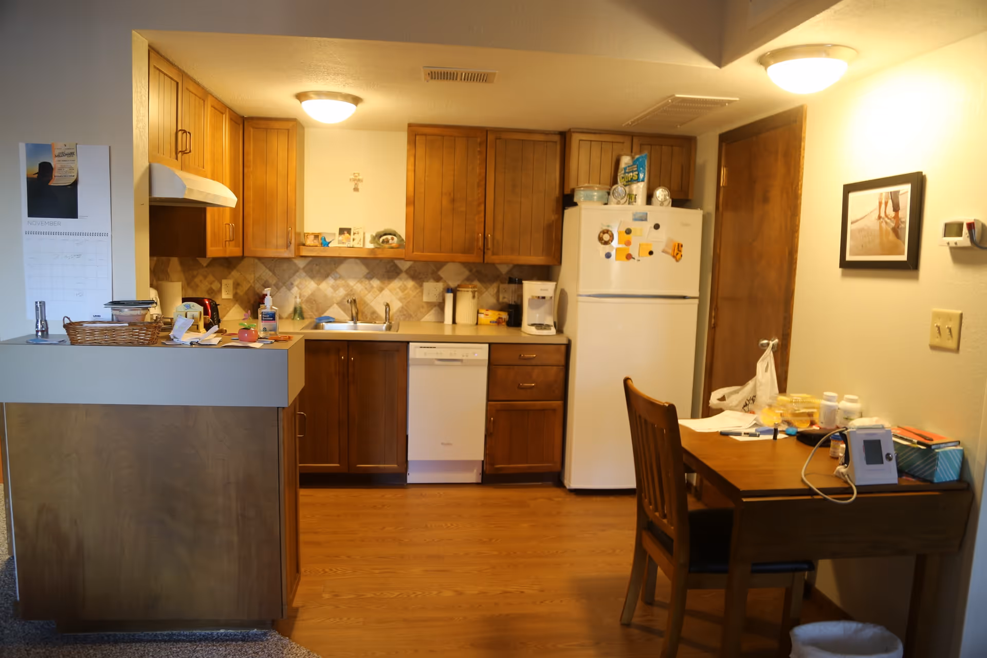A small kitchen area with wooden cabinets, a white refrigerator, a dishwasher, and a sink. There is a wooden table with a chair on the right side, holding various items including medication bottles and a medical device. The walls are light-colored with a framed picture and a calendar visible.