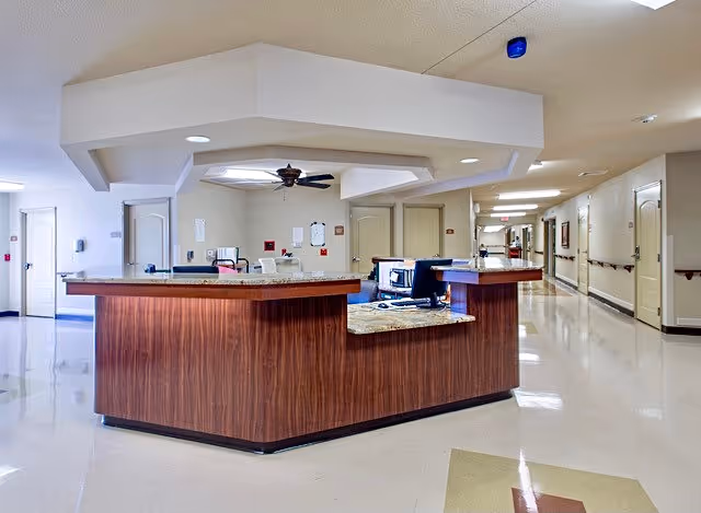 Interior view of a nursing and rehabilitation center reception area with a wooden front desk, computer monitors, and a ceiling fan. The hallway extends to the right with closed doors and handrails along the walls.