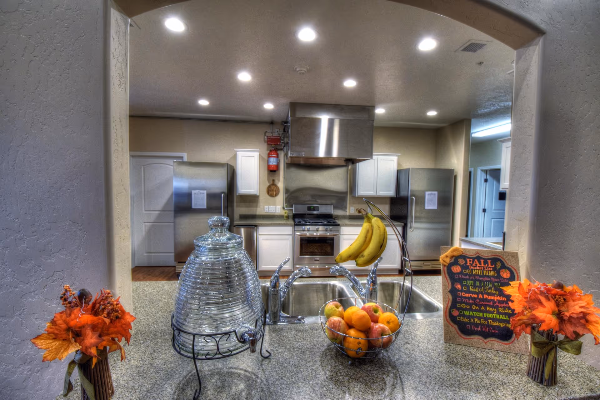 View of a modern kitchen with stainless steel appliances including two refrigerators and a stove with a vent hood. The kitchen has white cabinets and a granite countertop with a double sink. On the counter, there is a large glass water dispenser, a fruit basket with bananas, apples, and oranges, and two small autumn-themed floral arrangements. A decorative fall checklist sign is also displayed on the counter.