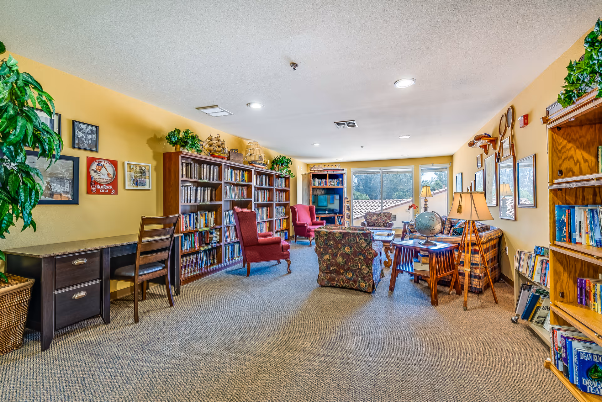 A cozy senior living facility common room with yellow walls, carpeted floor, and large windows letting in natural light. The room features multiple bookshelves filled with books, two red armchairs, a floral patterned armchair, a plaid sofa, a wooden desk with a chair, and various decorative items including plants, framed pictures, and model ships.
