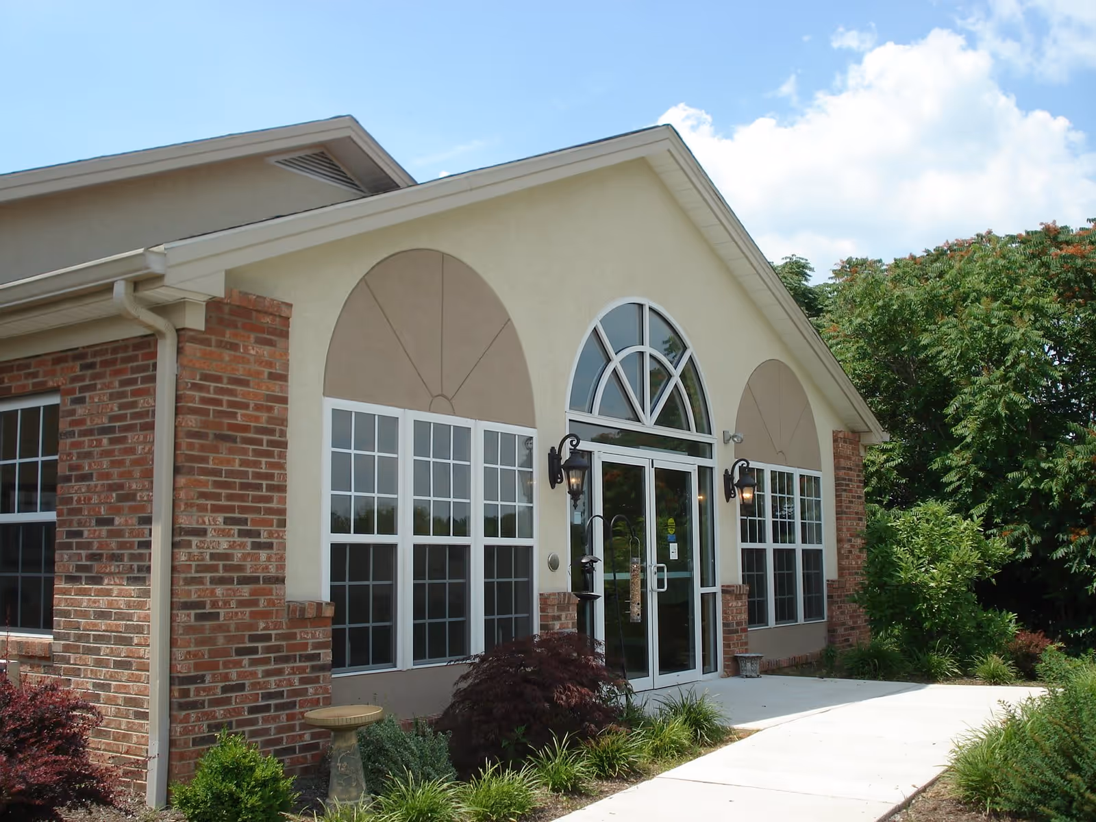 Exterior view of a single-story building with brick and beige stucco walls, large arched windows, and a glass double door entrance. The building is surrounded by greenery and a concrete walkway leading to the entrance under a partly cloudy sky.