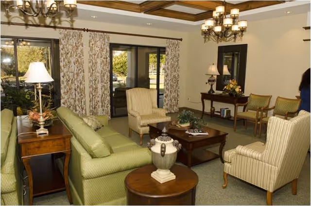 Bright furnished common living room with green sofas, striped armchairs, wooden coffee and side tables, chandeliers, and sliding glass doors to a patio.