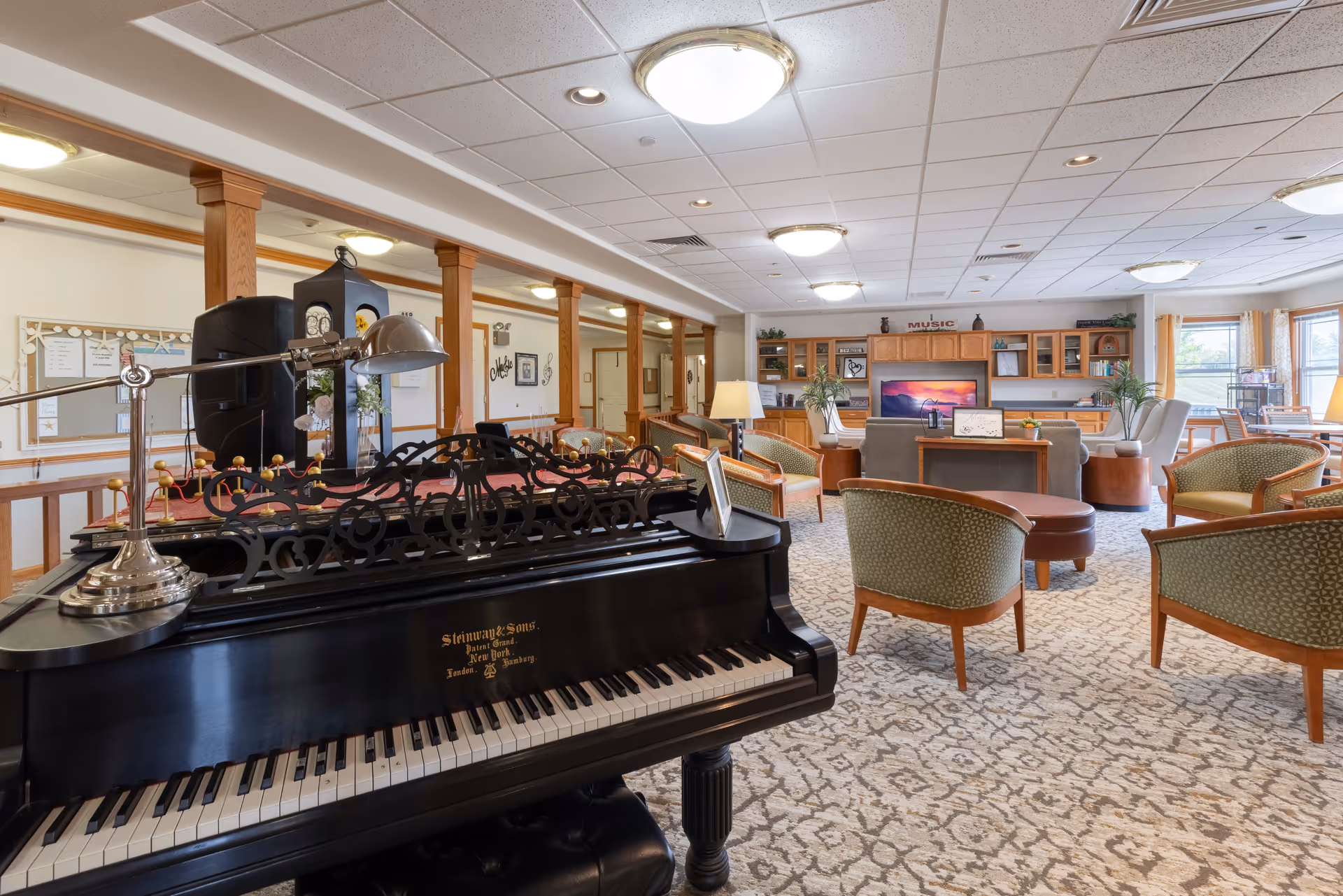 Spacious and well-lit common area in a senior living facility featuring a black grand piano in the foreground, several green upholstered chairs with wooden frames arranged around a central ottoman, a large flat-screen TV on a wooden cabinet, and large windows letting in natural light.