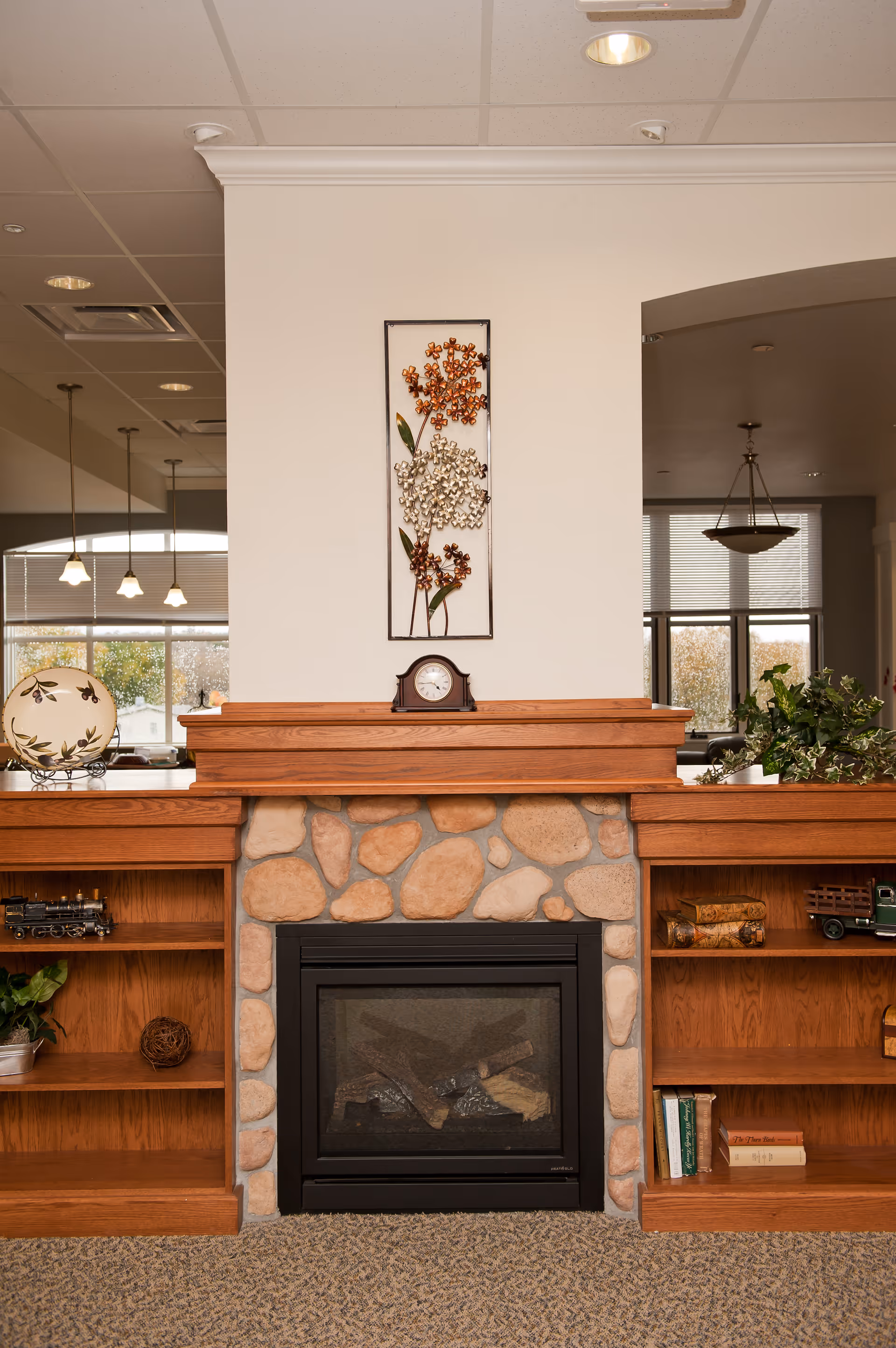 A cozy indoor fireplace with a stone surround and wooden mantel, flanked by built-in wooden shelves containing decorative items and books. Above the mantel is a wall decoration featuring metal flowers. The room has a carpeted floor and large windows in the background letting in natural light.