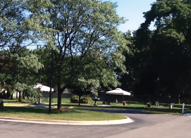 Outdoor area with green trees, grass, a paved road, and white umbrellas providing shade over tables in the background under a clear blue sky.