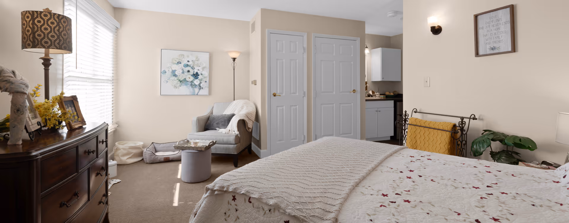 A cozy bedroom with a bed covered in a floral quilt and a beige knitted blanket. To the right of the bed is a decorative metal blanket holder with a yellow blanket and a green plant beside it. On the left side of the room, there is a wooden dresser with a lamp, framed photos, and yellow flowers. A comfortable armchair with a gray pillow and a white throw blanket sits near two closed white doors. A small round ottoman and a pet bed are on the carpeted floor. The walls are painted beige with a floral painting and a framed quote hanging on them. A glimpse of a kitchenette area is visible through an open doorway.