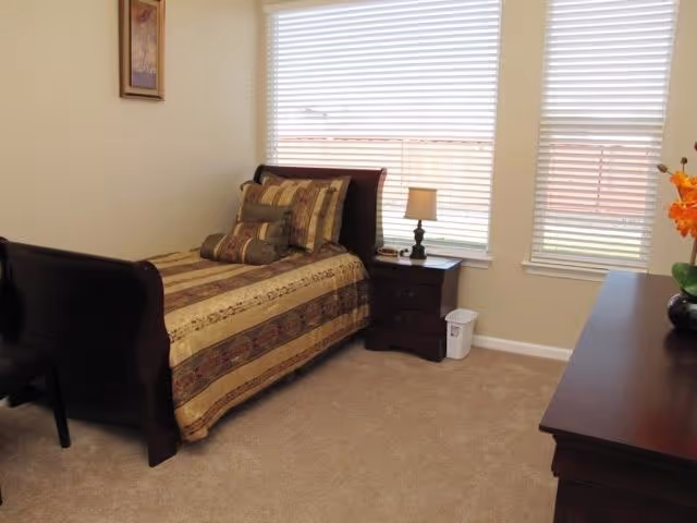 A cozy bedroom with a single bed featuring a brown and beige striped comforter and multiple pillows. Next to the bed is a dark wooden nightstand with a small lamp and a phone. The room has beige carpet and walls, two large windows with white blinds, a framed picture on the wall, and a dark wooden dresser with a vase of orange flowers on top.
