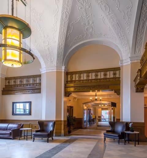 Spacious interior lobby area with high, ornately decorated ceilings and large archways. The room features leather armchairs and sofas arranged around small tables, with warm lighting from hanging fixtures. The walls have decorative molding and a framed mirror, and the floor is tiled with a patterned design. A hallway leads to a glass door entrance in the background.