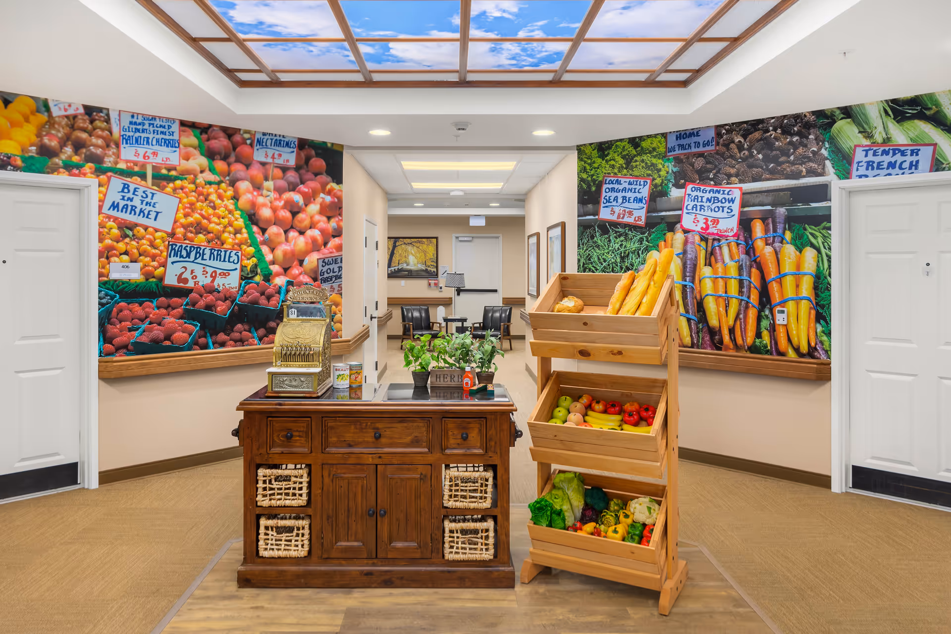 Interior hallway of a senior living facility with a wooden display stand holding artificial fruits and vegetables, a wooden cabinet with baskets and a vintage cash register on top, and large colorful murals of fresh produce on the walls. The ceiling features a skylight with a blue sky and clouds design. Two white doors are visible on either side of the hallway, and a seating area with chairs and a table is seen in the background.