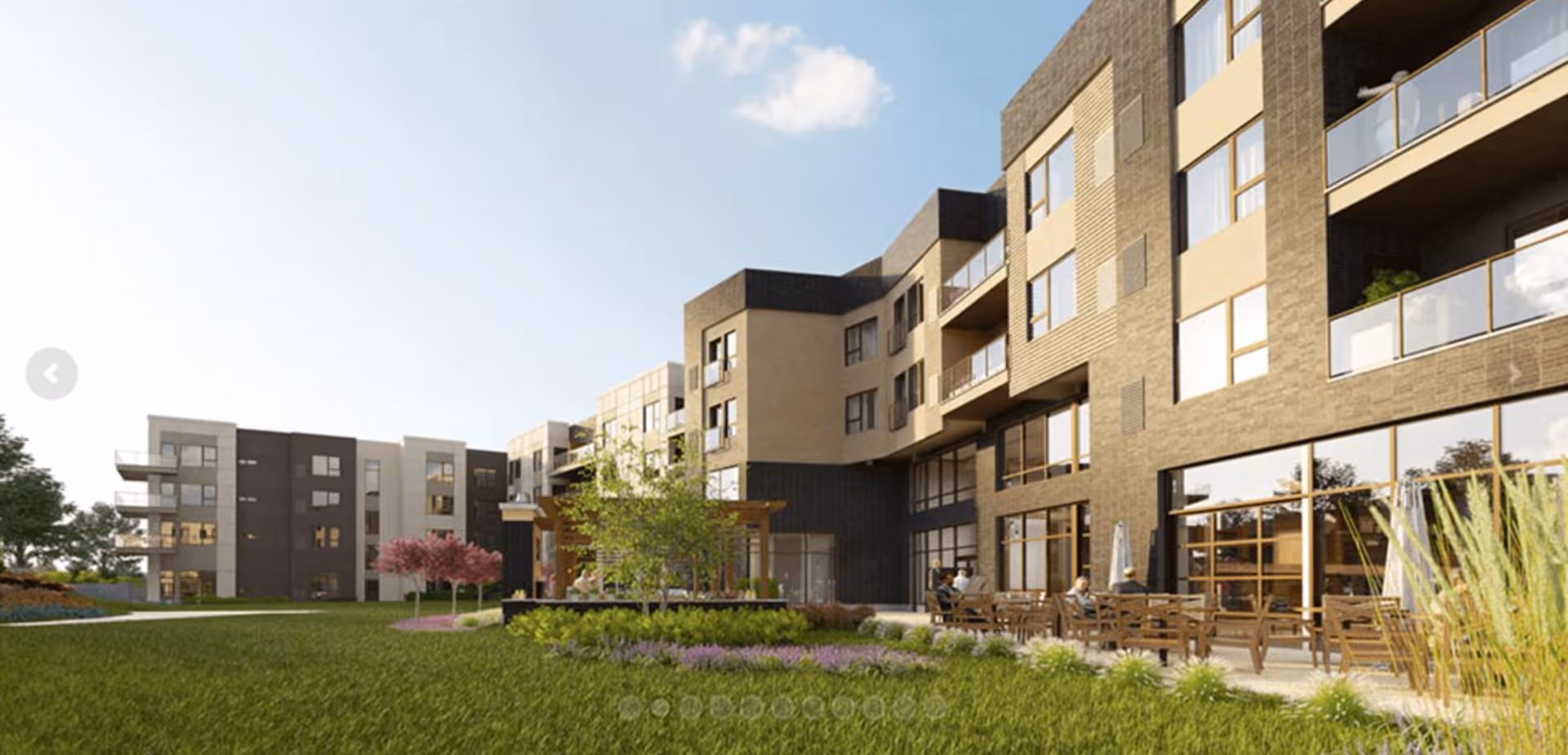 Outdoor view of a modern multi-story senior living facility with balconies, large windows, landscaped green lawn, trees, and a patio area with wooden tables and chairs where people are seated.