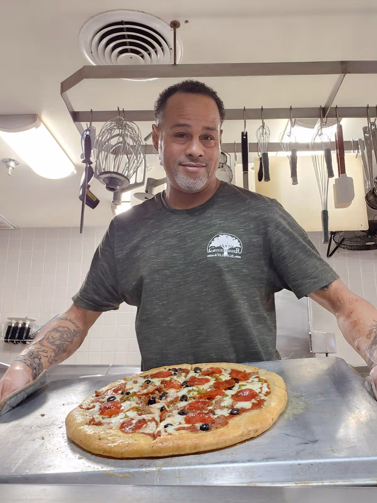 A man wearing a Greenbrier Village t-shirt is holding a large tray with a freshly baked pepperoni pizza topped with olives and green peppers in a kitchen setting with hanging kitchen utensils in the background.