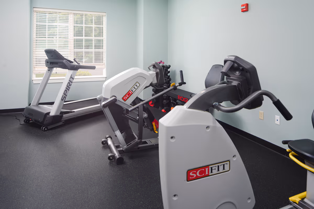 A small fitness room with exercise equipment including a treadmill, a recumbent bike, a rowing machine, and a rack of dumbbells. The room has light blue walls, a large window with blinds, and black rubber flooring.
