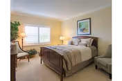 A cozy bedroom with a wooden bed frame, beige bedding, and multiple pillows. The room has light-colored walls and carpet, a window with white shutters, a framed picture above the bed, a floor lamp, a table lamp on a nightstand, a wooden chair with a cushion, a small plant, and an upholstered armchair.