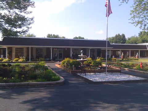 Front exterior view of Curis Waynesboro Nursing and Rehabilitation Center showing a single-story building with a covered entrance, landscaped garden with a fountain, benches, and an American flag on a flagpole in front.