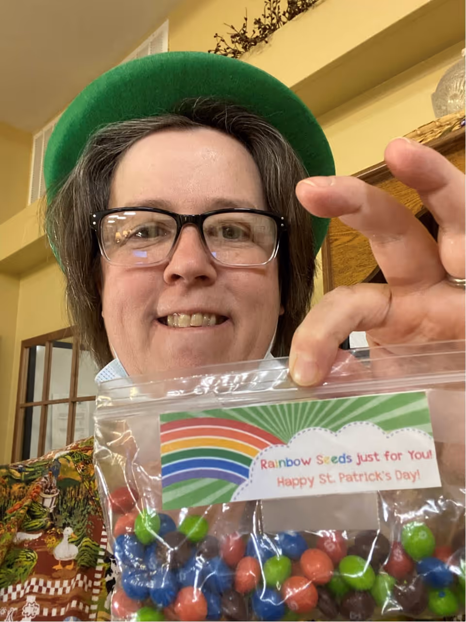 A person wearing glasses and a green hat smiles while holding a clear plastic bag filled with colorful candy. The bag has a label with a rainbow and the text 'Rainbow Seeds just for You! Happy St. Patrick's Day!'. The background shows an indoor setting with yellow walls and wooden furniture.
