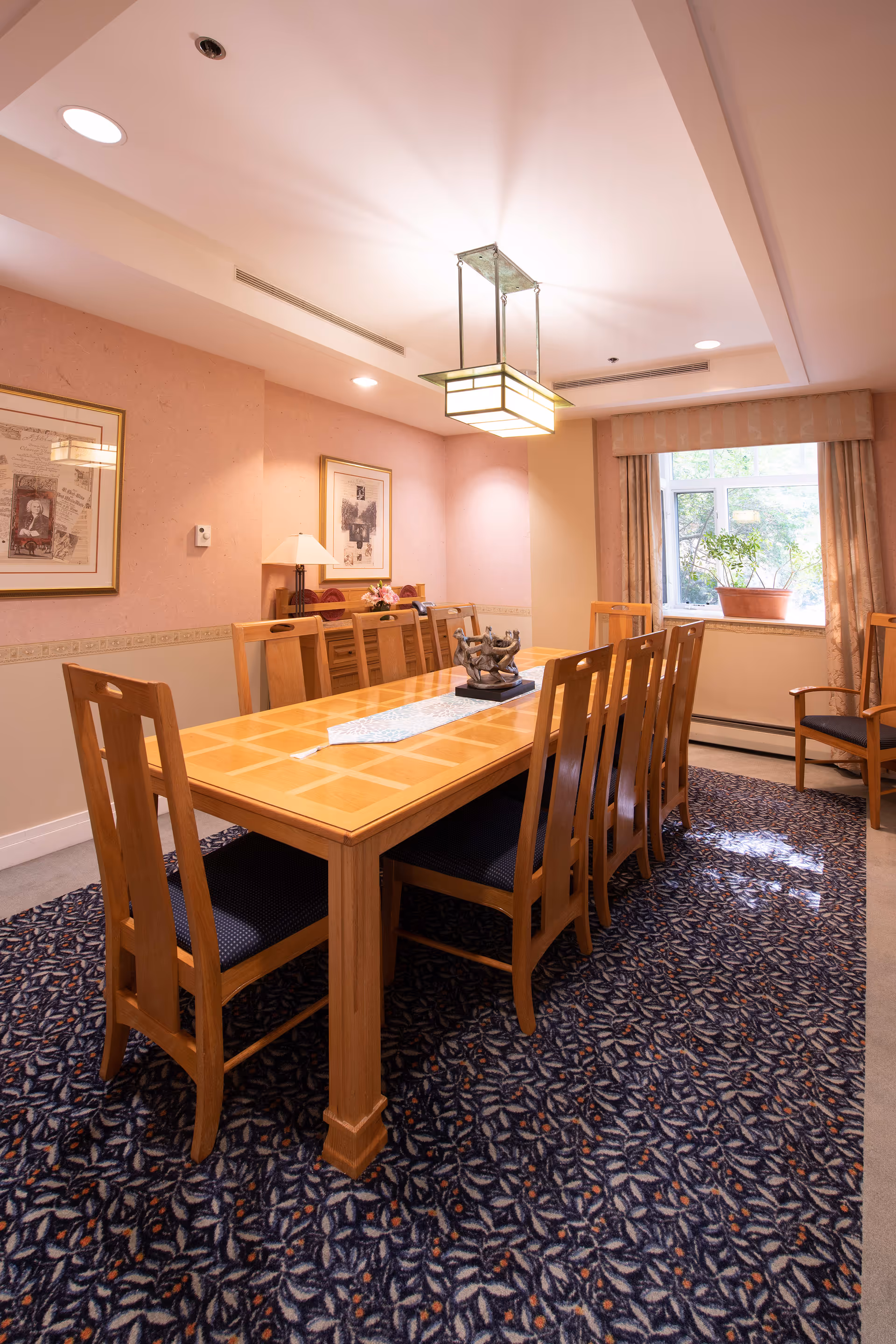 A long wooden dining table surrounded by chairs under a pendant light in a carpeted dining room with framed art and a window.