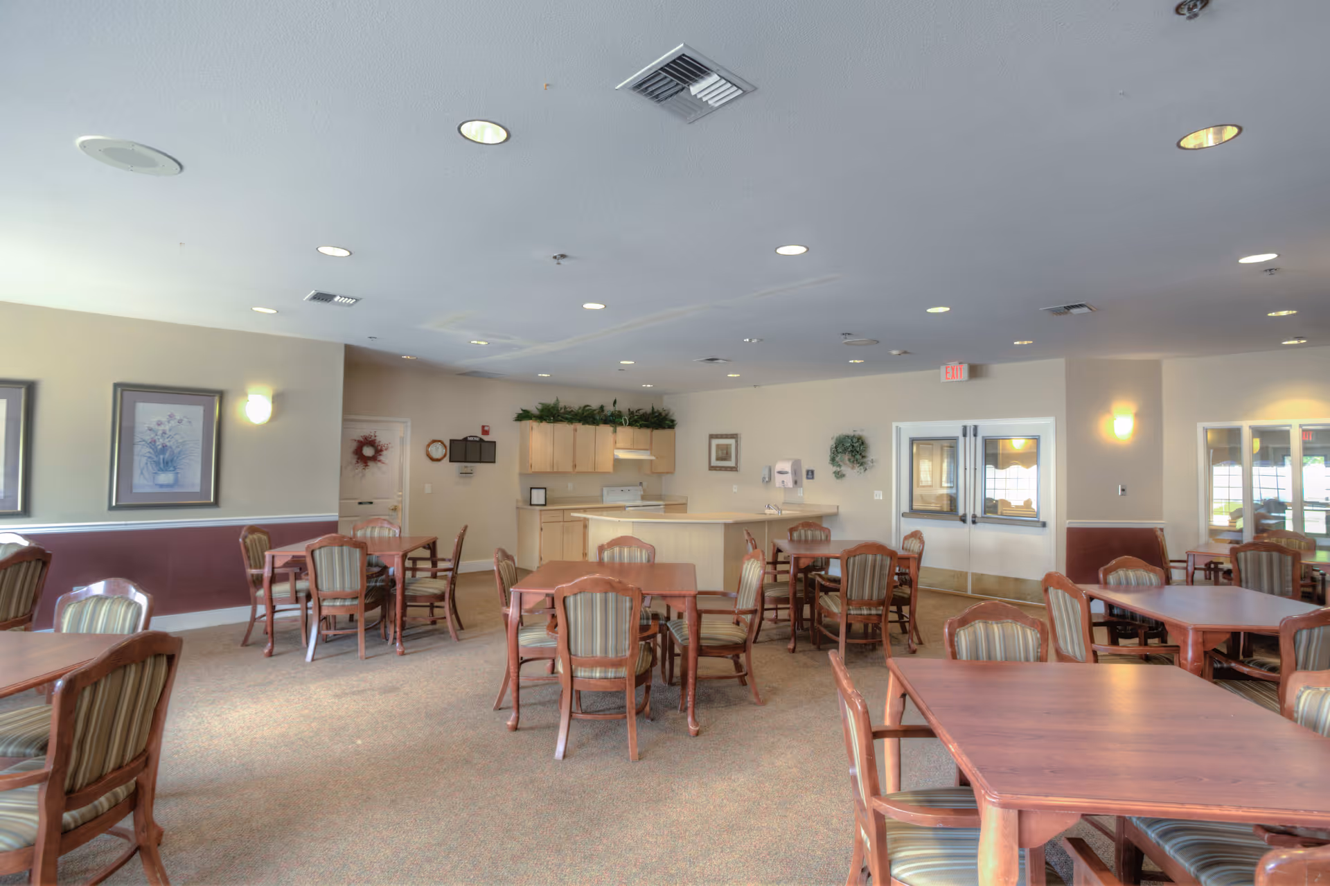A spacious dining room with multiple wooden tables and chairs arranged neatly. The room has beige walls with maroon wainscoting, framed floral artwork, and soft lighting from wall sconces and ceiling lights. In the background, there is a small kitchen area with light wood cabinets, a stove, and a countertop. Double doors and windows are visible on the right side of the room.