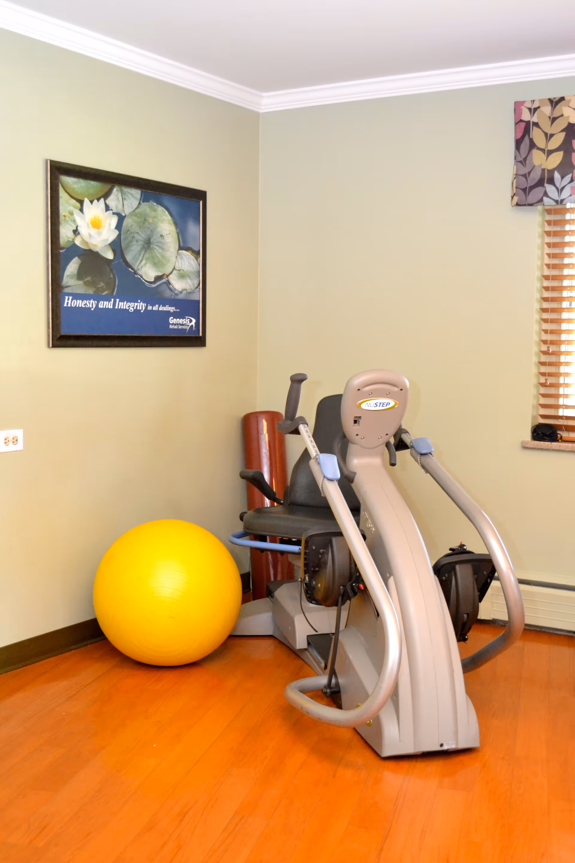 A small exercise room with a recumbent cross trainer machine, a large yellow exercise ball, and a foam roller against a beige wall. There is a framed poster on the wall with a water lily and the text 'Honesty and Integrity in all dealings... Genesis Rehab Services'. A window with wooden blinds and a floral valance is partially visible on the right.
