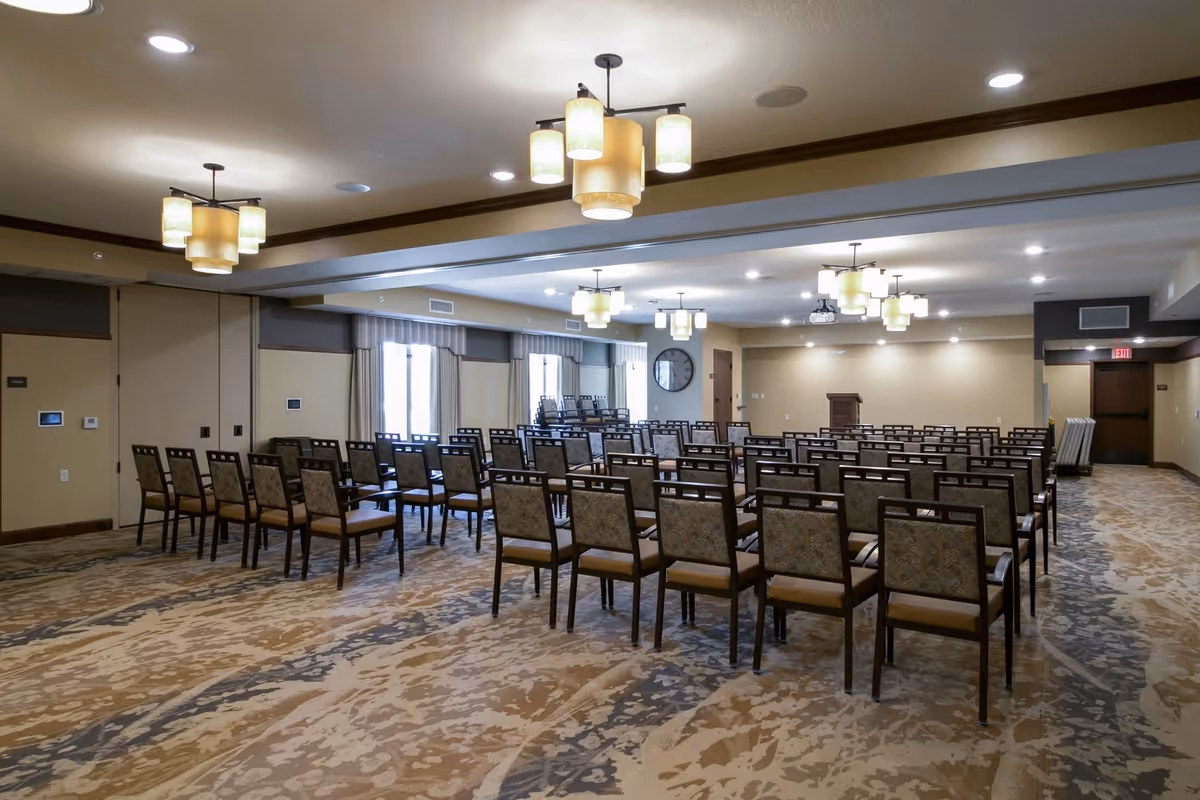A large conference or meeting room with rows of chairs arranged facing a wooden podium at the front. The room has patterned carpet flooring, beige walls, multiple ceiling light fixtures, and windows with curtains along one side.