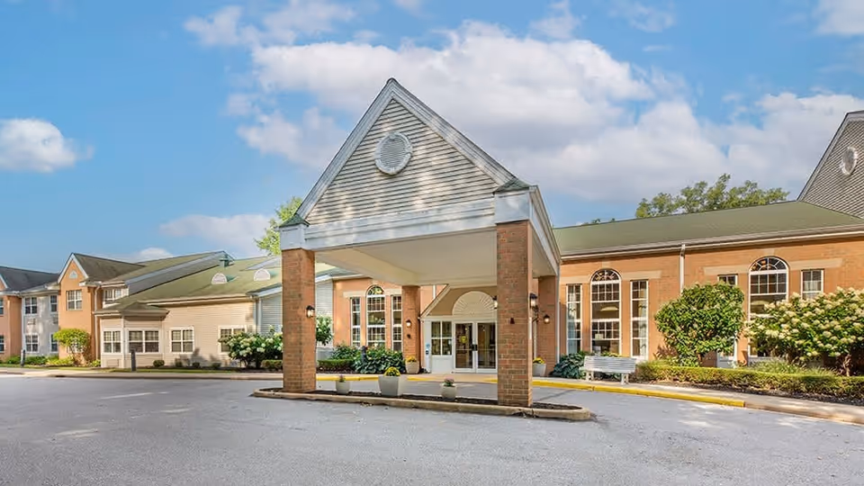 Exterior view of Brookdale Wickliffe senior living facility showing the main entrance with a covered drop-off area supported by brick columns, surrounded by well-maintained landscaping and a clear blue sky with some clouds.