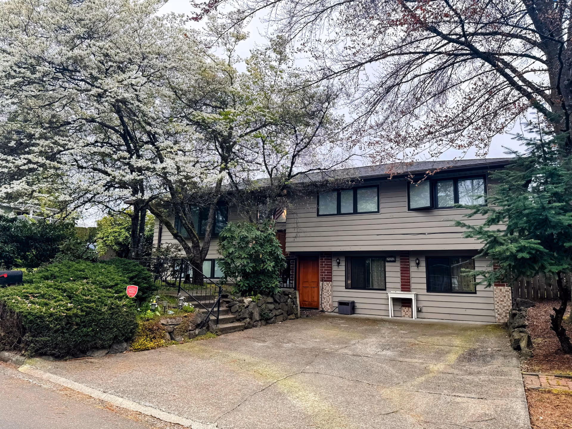 Exterior view of a two-story residential building with beige siding and brick accents, surrounded by trees with white blossoms and green shrubs. There is a concrete driveway in front of the building and a wooden front door with steps leading up to it.