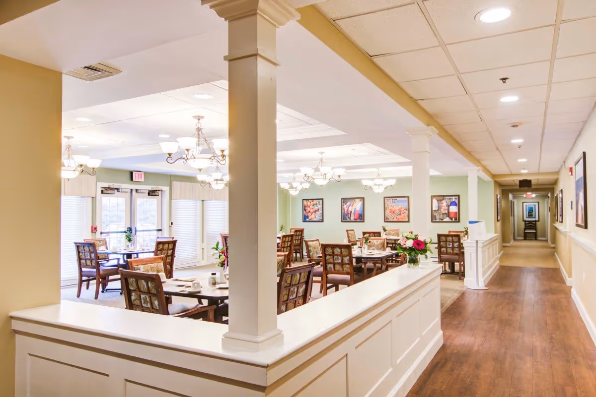 A bright and spacious dining room in a senior living facility with multiple wooden tables and chairs arranged neatly. The room features soft green walls adorned with framed artwork, elegant chandeliers hanging from a white coffered ceiling, and large windows with white blinds allowing natural light to fill the space. A long white half-wall with columns separates the dining area from a hallway with wood flooring and additional framed pictures on the walls.