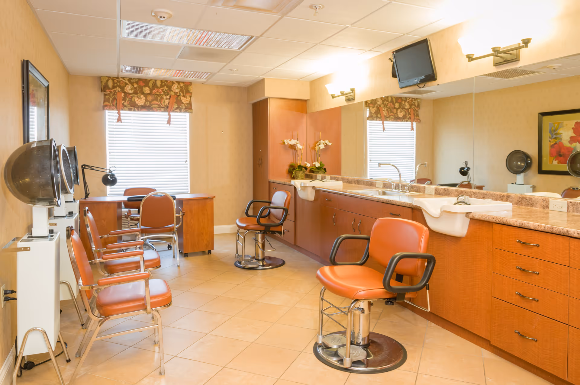 A bright salon room with several orange salon chairs, hair dryers, a large mirror above a countertop with sinks, a small desk with a chair near two windows with floral valances, and a wall-mounted TV.